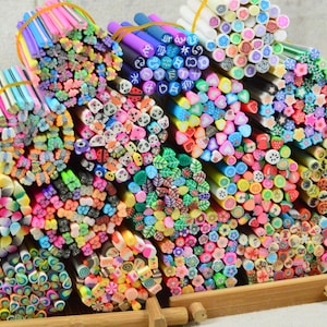 Puede incluir: Una colección de cañas de arcilla polimérica coloridas, agrupadas con bandas elásticas. Las cañas muestran varios diseños, incluyendo caras sonrientes, flores, frutas y otros patrones decorativos. Los colores vibrantes y los detalles intrincados son ideales para manualidades.