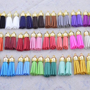 May include: Assortment of 36 colorful suede tassels with gold metal caps. The tassels are arranged in rows of two, with each row featuring a different color. The colors include red, orange, yellow, green, blue, purple, pink, brown, white, and gray.