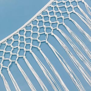 May include: A white macrame fringe trim featuring a woven header with a diamond-shaped knotted net pattern. The trim extends into long, straight, vertical hanging cords that create a decorative edge for textile projects and home decor items.