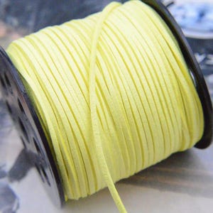 May include: A spool of light yellow cord wrapped around a black plastic reel. The cord has a flat, suede-like texture and is wound tightly in multiple parallel rows across the entire width of the black spool, with one loose end extending outward.