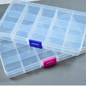 Può includere: Due scatole trasparenti in plastica con più scomparti sono impilate. Una scatola ha una chiusura blu, l'altra rosa. Le scatole sono su una superficie grigia.