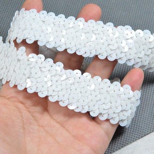 May include: Close-up of a white sequin trim. The trim is made of small, round, white sequins sewn onto a fabric backing. The trim is held in a hand, showing its texture and detail. Ideal for embellishing clothing or crafts.