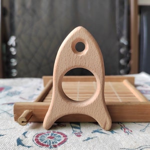 May include: A wooden rocket toy with a smooth, light brown finish. The rocket has a large, round opening in the middle and a small hole at the top.