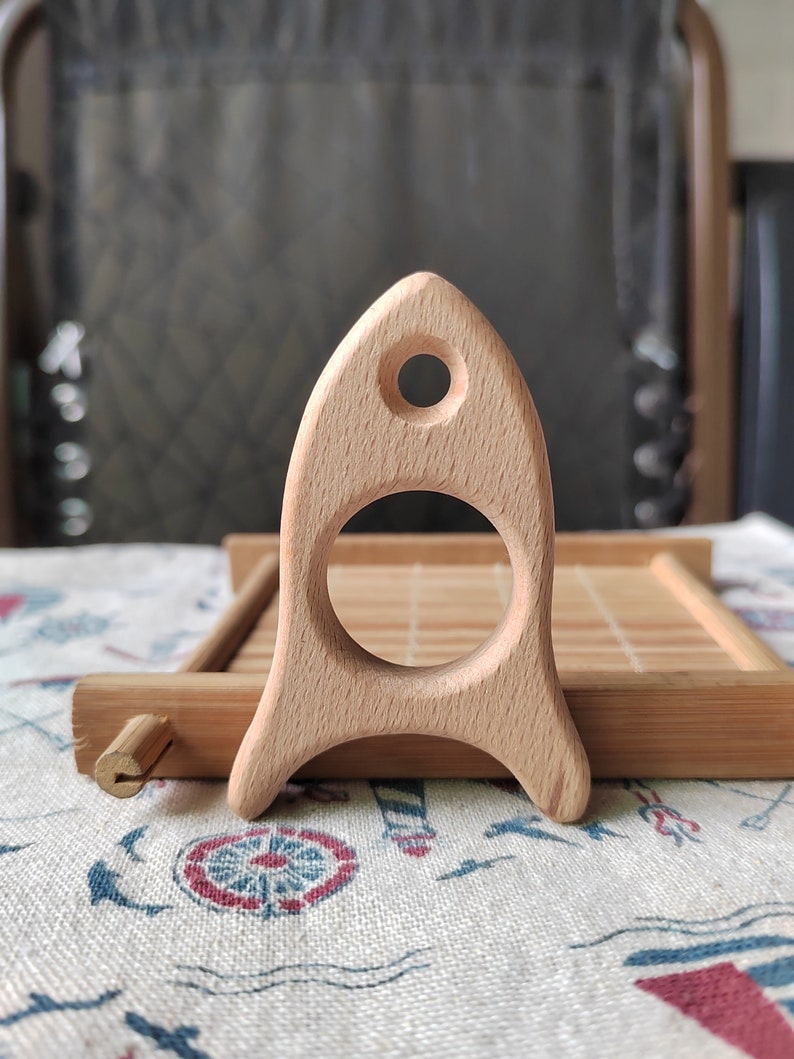 May include: A wooden rocket toy with a smooth, light brown finish. The rocket has a large, round opening in the middle and a small hole at the top.