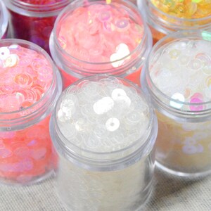 May include: Close-up of several small clear jars filled with colorful sequins. The sequins are in shades of pink, white, yellow, and purple. The jars are arranged closely together, with their clear lids visible.