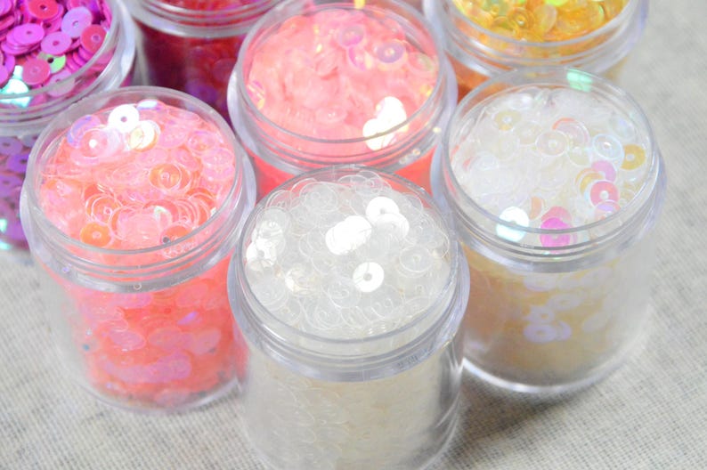 May include: Close-up of several small clear jars filled with colorful sequins. The sequins are in shades of pink, white, yellow, and purple. The jars are arranged closely together, with their clear lids visible.