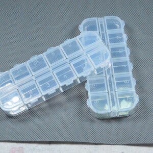 Peut inclure: Deux piluliers en plastique transparent avec plusieurs compartiments, chacun num&eacute;rot&eacute;. Les piluliers sont rectangulaires et dot&eacute;s d'une fermeture &agrave; pression. Ils sont pos&eacute;s sur une surface grise textur&eacute;e.