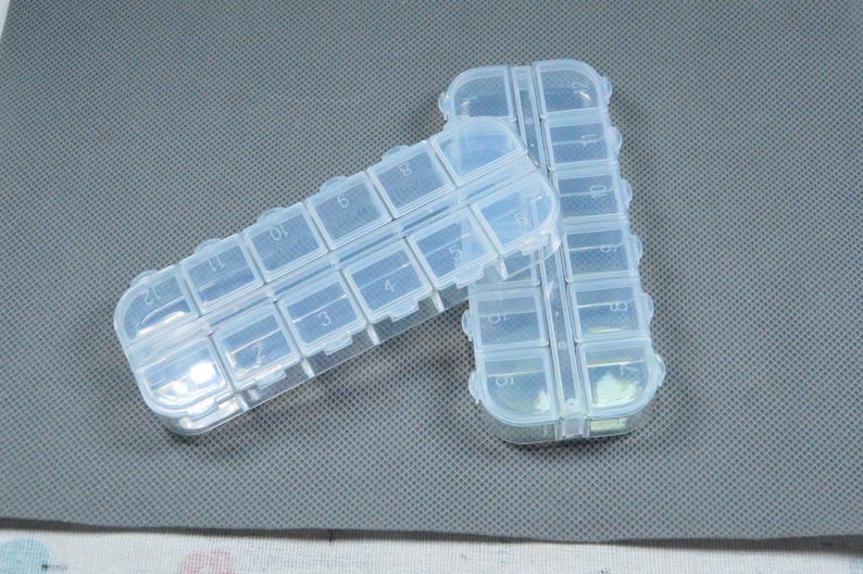Peut inclure: Deux piluliers en plastique transparent avec plusieurs compartiments, chacun num&eacute;rot&eacute;. Les piluliers sont rectangulaires et dot&eacute;s d'une fermeture &agrave; pression. Ils sont pos&eacute;s sur une surface grise textur&eacute;e.