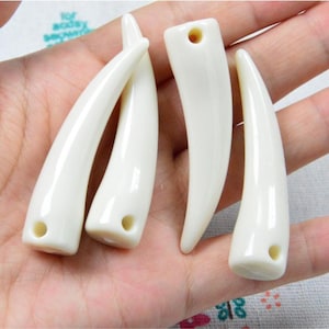 May include: Four off-white, curved pendants resembling fangs. Each has a smooth, glossy finish and a small hole drilled near the base. The pendants are displayed against a neutral background, highlighting their shape and color.
