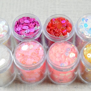 May include: Eight small, clear plastic jars filled with colorful sequins. The sequins are in various colors, including white, pink, red, blue, and gold. The jars are arranged in two rows of four.