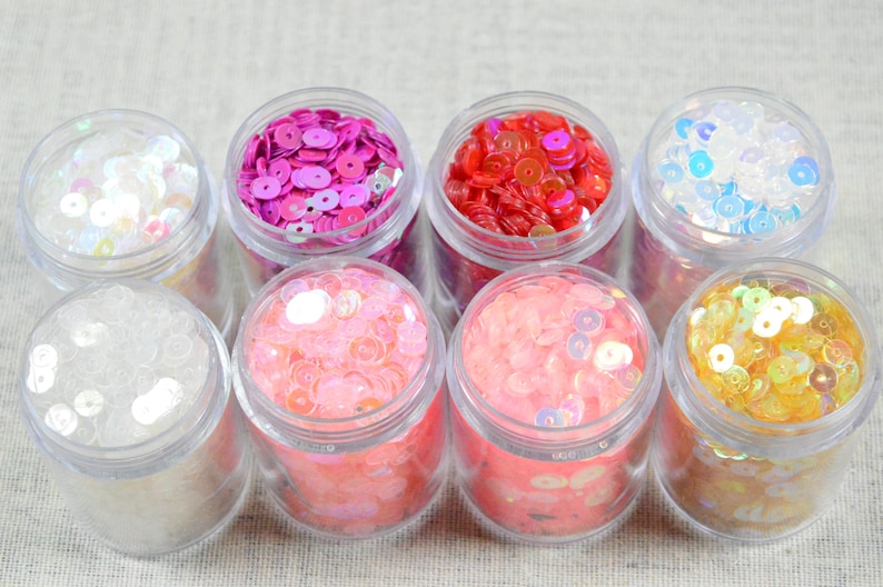 May include: Eight small, clear plastic jars filled with colorful sequins. The sequins are in various colors, including white, pink, red, blue, and gold. The jars are arranged in two rows of four.