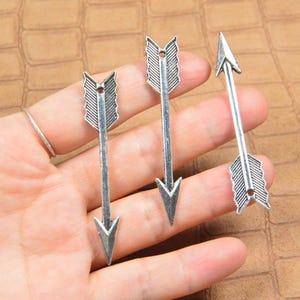 May include: Three silver-toned metal arrow charms featuring a textured fletching design and a small circular hole at the top for attachment. Each arrow has a pointed tip and a detailed shaft, presented as individual components for jewellery making.