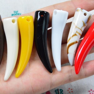 Puede incluir: Una colección de siete colgantes curvos en forma de cuerno en varios colores, incluyendo marrón, crema, amarillo, negro, blanco, blanco con vetas marrones y rojo. Cada colgante presenta un acabado liso y brillante con un pequeño orificio superior.