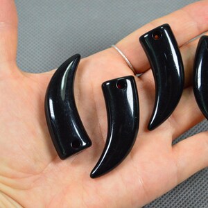 Puede incluir: Cuatro colgantes negros lisos y pulidos con forma de dientes o garras de animal curvadas. Cada colgante presenta un pequeño orificio circular en la parte superior para enhebrar, mostrando una superficie brillante y un color negro sólido.
