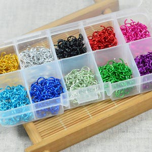 Puede incluir: Una caja organizadora de plástico transparente llena de anillas de colores. Las anillas son de varios colores, incluyendo dorado, plateado, negro, rojo, rosa, morado, azul, verde y turquesa. La caja está sobre una bandeja de madera.