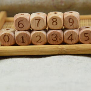 Puede incluir: Bloques de números de madera del 0 al 9, dispuestos en dos filas sobre una pequeña bandeja de bambú. Cada bloque es un cubo marrón claro con un número grabado en una fuente marrón más oscura. La bandeja también es de bambú.