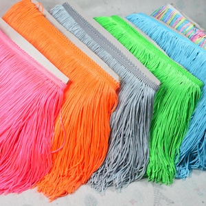 May include: A collection of six rolls of fringe trim featuring vibrant colours including neon pink, bright orange, light grey, lime green, sky blue, and a multi-coloured rainbow pattern. Each roll consists of long, thin, hanging fabric strands.