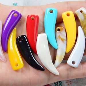 Puede incluir: Una colección de once colgantes curvos en forma de diente en varios colores y acabados, incluyendo rojo, amarillo, negro, blanco y turquesa sólidos, junto con patrones jaspeados en rosa, marrón y crema, cada uno con un orificio perforado.