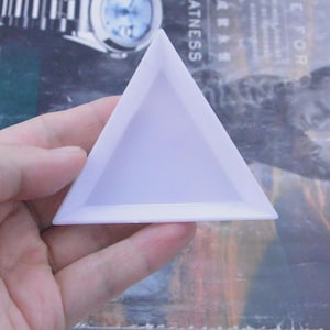 Peut inclure: Un plateau triangulaire en plastique blanc avec une base plate et des bords surélevés. Le plateau présente une finition lisse et mate ainsi qu'une forme géométrique simple, conçue pour contenir de petits objets ou des perles.