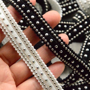 Puede incluir: Dos ribetes de tela negra y blanca con cuentas plateadas cosidas. Los ribetes se sostienen en una mano.
