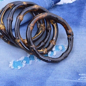 Op de afbeelding: Zes donkerbruine bamboe ringen met een glanzende afwerking en zichtbare bamboenervaring. De ringen liggen op een blauwe denim stof achtergrond.