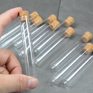 Pode incluir: Tubos de ensaio de vidro transparente com rolhas de cortiça. Um tubo é segurado por uma mão, com outros ao fundo. As rolhas são de cor castanho claro, os tubos são transparentes.