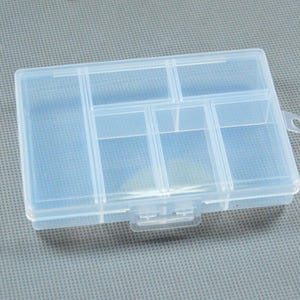 May include: A clear, rectangular plastic organiser box with multiple compartments. The box has a hinged lid and a small loop on one side. The compartments are divided by vertical and horizontal dividers.