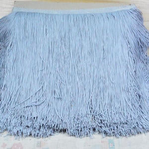 May include: A pale blue fringe trim with a beige backing. The fringe is composed of numerous fine threads, giving a soft, textured look. This trim is suitable for various craft and design projects, such as clothing or home decor.