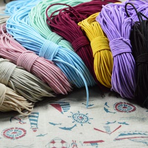 Puede incluir: Una colección de cordones elásticos coloridos en varios tonos, incluyendo beige, rosa, azul, burdeos, amarillo, morado y negro. Los cordones están agrupados y atados, mostrando una gama de opciones para manualidades o proyectos de bricolaje. Los cordones están sobre una tela de temática náutica.
