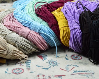 May include: A collection of colorful elastic cords in various hues, including beige, pink, blue, burgundy, yellow, purple, and black. The cords are bundled and tied, showcasing a range of options for crafting or DIY projects. The cords are on a nautical-themed fabric.