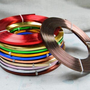 May include: A stack of colorful metallic strips in various colors, including red, gold, green, blue, and pink. A separate coil of copper-colored strips rests nearby. The strips are bound with white string, suggesting they are for crafting or decorative purposes.