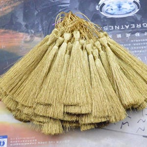May include: A collection of gold-coloured silk tassels, each featuring a thin matching cord loop at the top. The tassels are arranged in a fan-like cluster, showcasing the fine, straight, and shimmering threads that form the base of each individual piece.