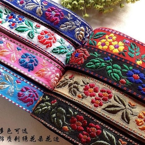 以下が含まれることがあります： 青、ピンク、赤、黒など、さまざまな色の刺繍された花のリボンのクローズアップ。リボンは、緑の葉とカラフルな花で詳細な花のパターンを特徴としています。画像内のテキストは「複数の色をご利用いただけます、綿刺繍の花柄レースエッジ」と表示されています。