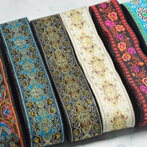 Könnte beinhalten: Sechs Streifen aus dekorativem Webband, die nebeneinander angeordnet sind. Die Bänder weisen komplexe geometrische und florale Muster in leuchtenden Farben wie Gold, Blau, Rot, Pink und Schwarz auf, durchzogen von eingewebten Metallic-Fäden.