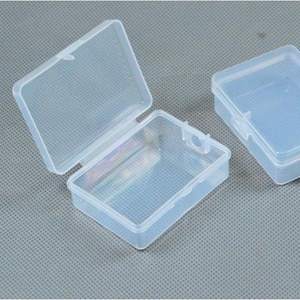 May include: Three clear, rectangular plastic storage boxes. One box is open, revealing the interior, while the other two are closed. The boxes have a hinged lid and a small latch. The boxes are on a gray surface.