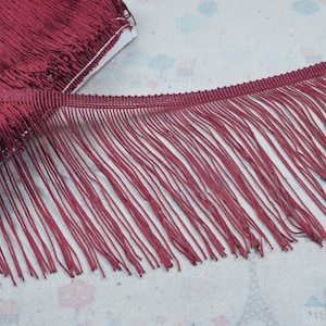 Puede incluir: Un carrete de flecos de color burdeos intenso con una cinta de cabecera tejida y hilos largos, finos y colgantes. El fleco está densamente empaquetado y es uniforme en color, mostrando una textura consistente en toda la longitud.