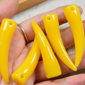 May include: Five smooth, bright yellow horn-shaped pendants, each featuring a small circular hole at the top for stringing. These curved, tapered charms have a glossy finish and a solid, uniform colour throughout their entire structure.