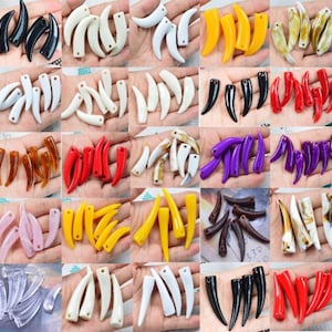 Puede incluir: Una colección de colgantes con forma de diente de varios colores. Los colgantes son de color negro, blanco, amarillo, rojo, marrón, morado y transparente. Cada colgante tiene un agujero para ensartar. Los colgantes se muestran en varias manos.
