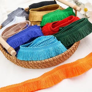 May include: A selection of colourful fringe trim rolls displayed in a woven basket. The trims include shades of blue, green, red, gold, and black. An orange fringe trim is in the foreground. These trims are likely for sewing, crafting, or decorative use.