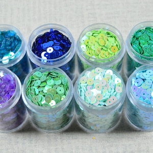 May include: Eight small, clear plastic jars filled with colorful sequins. The sequins are in shades of blue, green, purple, and iridescent white. The jars are arranged in two rows on a neutral surface.
