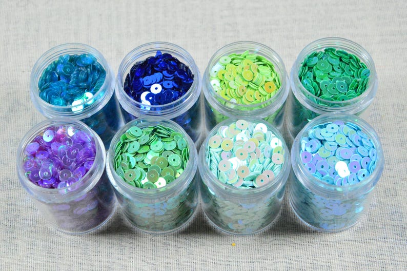 May include: Eight small, clear plastic jars filled with colorful sequins. The sequins are in shades of blue, green, purple, and iridescent white. The jars are arranged in two rows on a neutral surface.