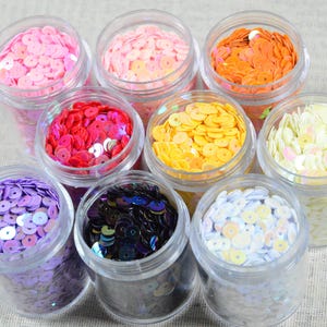 May include: An overhead shot displays eight clear plastic jars filled with colorful sequins. The sequins are in shades of pink, orange, red, yellow, purple, black, and white. The jars are arranged on a neutral-colored fabric surface.