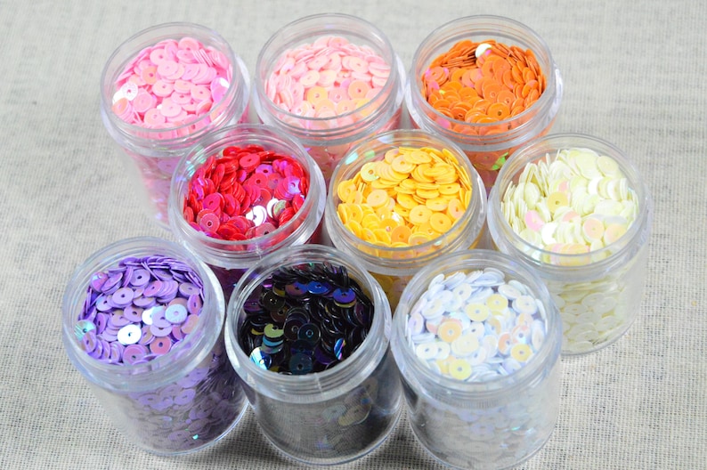 May include: An overhead shot displays eight clear plastic jars filled with colorful sequins. The sequins are in shades of pink, orange, red, yellow, purple, black, and white. The jars are arranged on a neutral-colored fabric surface.