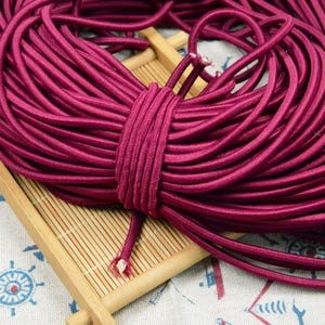May include: A bundle of burgundy-colored elastic cord, tightly wound and secured. The cord is smooth and appears to be made of a synthetic material. The bundle rests on a small, light-colored wooden tray, with a nautical-themed patterned fabric in the background.