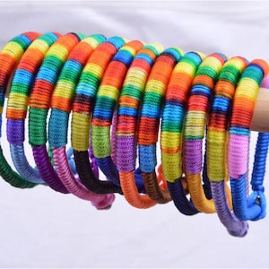 Puede incluir: Una colección de pulseras de colores del arcoíris. Cada pulsera está envuelta en hilos vibrantes de rojo, naranja, amarillo, verde, azul y morado. Las pulseras se exhiben en una clavija de madera sobre un fondo blanco.