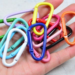 May include: A collection of small, oval-shaped metal carabiner clips in a variety of vibrant colors including yellow, red, blue, green, orange, white, black, pink, and light purple. Each carabiner features a spring-loaded gate mechanism.