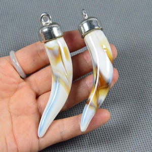 May include: Two pendant charms shaped like curved horns. The charms are white with streaks of yellow and brown, and have silver-colored metal caps and loops for hanging. The background is a gray surface.