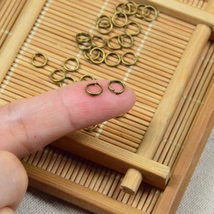 May include: A close-up of a finger holding two small, gold-coloured jump rings. The jump rings are on a bamboo mat with other jump rings scattered around.