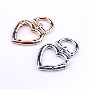 Könnte beinhalten: Zwei herzförmige Drehverschlüsse. Einer ist goldfarben, der andere silberfarben. Jeder Verschluss hat einen D-Ring und einen federbelasteten Verschluss. Die Verschlüsse sind zum Befestigen an Taschen, Schlüsselanhängern oder anderem Zubehör gedacht.