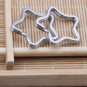 May include: Two silver-colored star-shaped keychains. The keychains are interlocked and have a wavy design. The keychains are made of metal and are on a light brown bamboo surface.